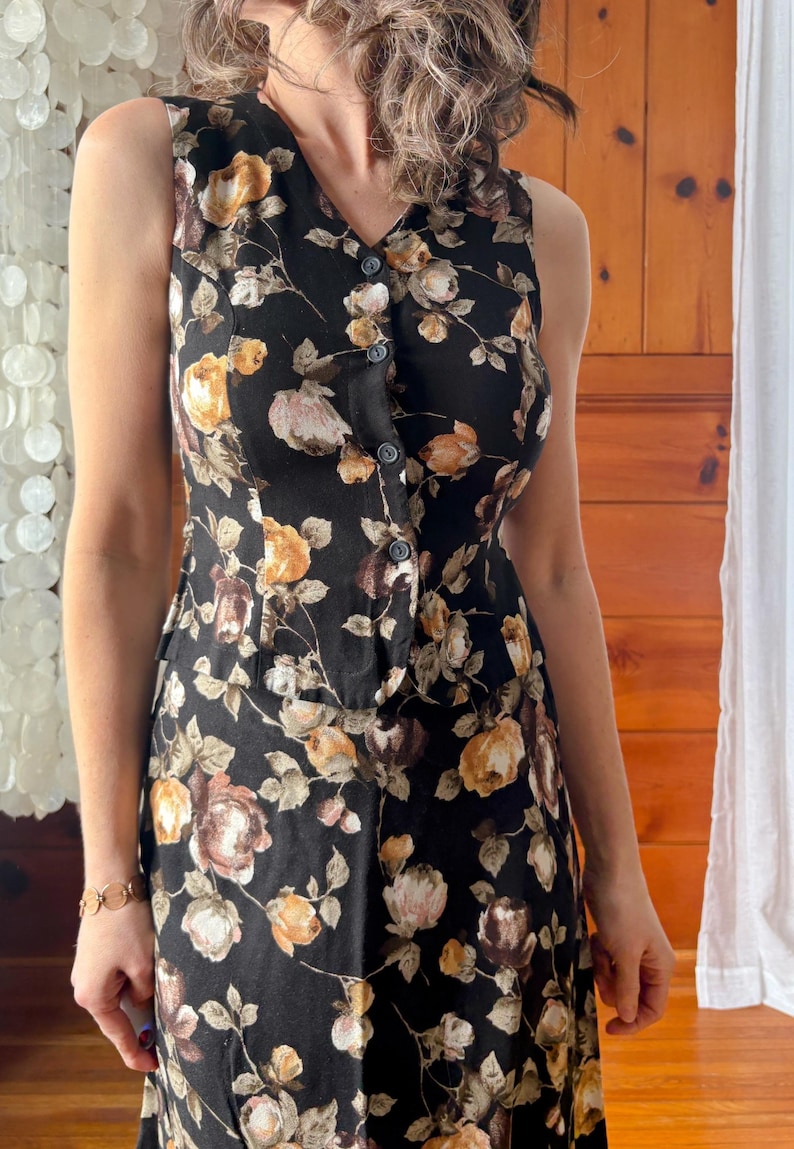 K&ouml;nnte beinhalten: Ein &auml;rmelloses schwarzes Kleid mit einem Blumendruck mit braunen, gelben und rosa Rosen. Das Kleid hat einen Knopfleisten vorne und ein tailliertes Oberteil.