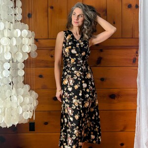 K&ouml;nnte beinhalten: Eine Frau tr&auml;gt ein schwarzes, florales Midikleid mit Knopfleiste und V-Ausschnitt. Sie steht vor einer Holzvert&auml;felung und tr&auml;gt nude High Heels.