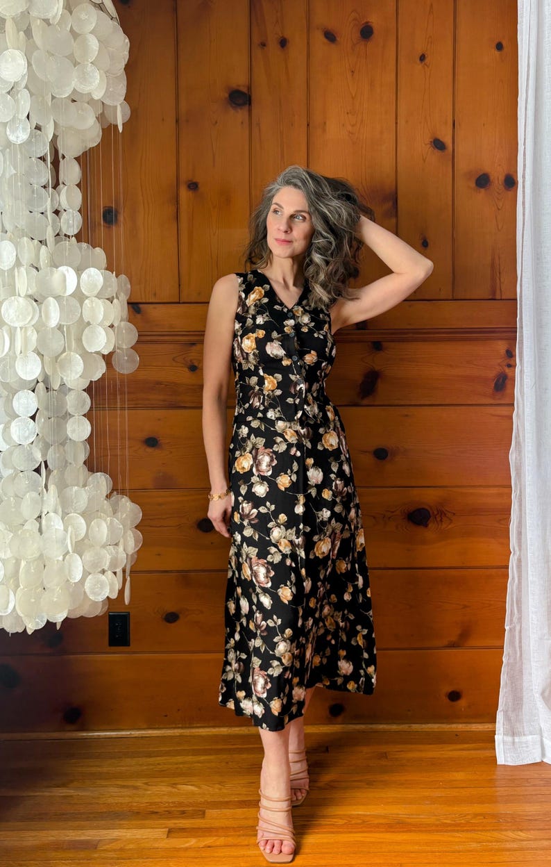 K&ouml;nnte beinhalten: Eine Frau tr&auml;gt ein schwarzes, florales Midikleid mit Knopfleiste und V-Ausschnitt. Sie steht vor einer Holzvert&auml;felung und tr&auml;gt nude High Heels.