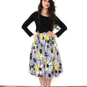 May include: A woman wearing a black long-sleeved shirt and a yellow, black, and white floral skirt. She is standing on a wooden floor.