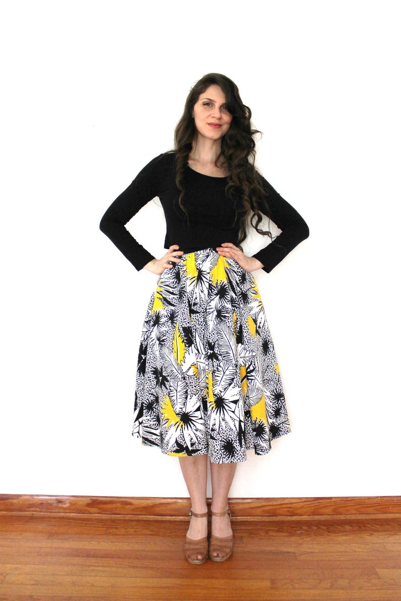 May include: A woman wearing a black long-sleeved shirt and a yellow, black, and white floral skirt. She is standing on a wooden floor.