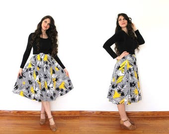 May include: A woman wearing a black long-sleeved shirt and a yellow, black, and white floral skirt. She is standing on a wooden floor with a white wall behind her.