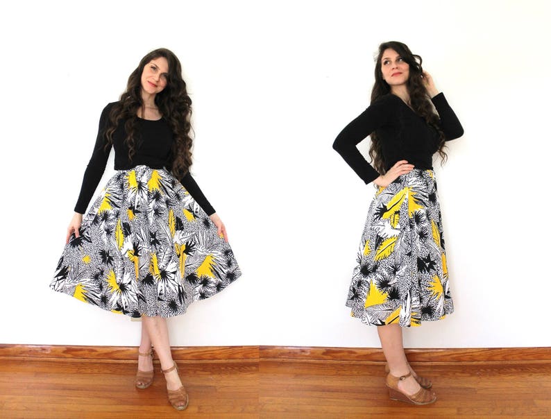 May include: A woman wearing a black long-sleeved shirt and a yellow, black, and white floral skirt. She is standing on a wooden floor with a white wall behind her.