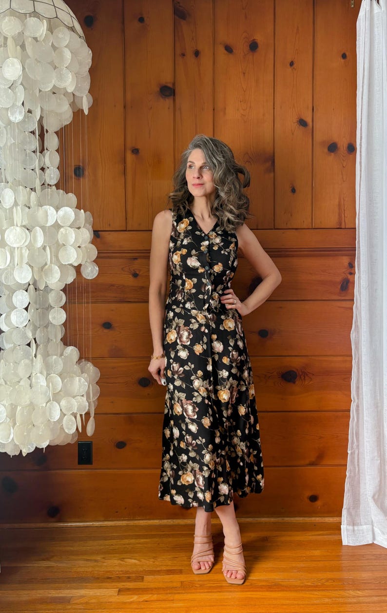K&ouml;nnte beinhalten: Eine Frau tr&auml;gt ein schwarzes Midikleid mit Blumenmuster und braunen Schuhen mit Absatz. Das Kleid hat einen V-Ausschnitt und eine Knopfleiste vorne.