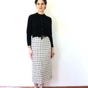 May include: A woman wearing a black cardigan and a white and black plaid skirt. She is wearing black heels.