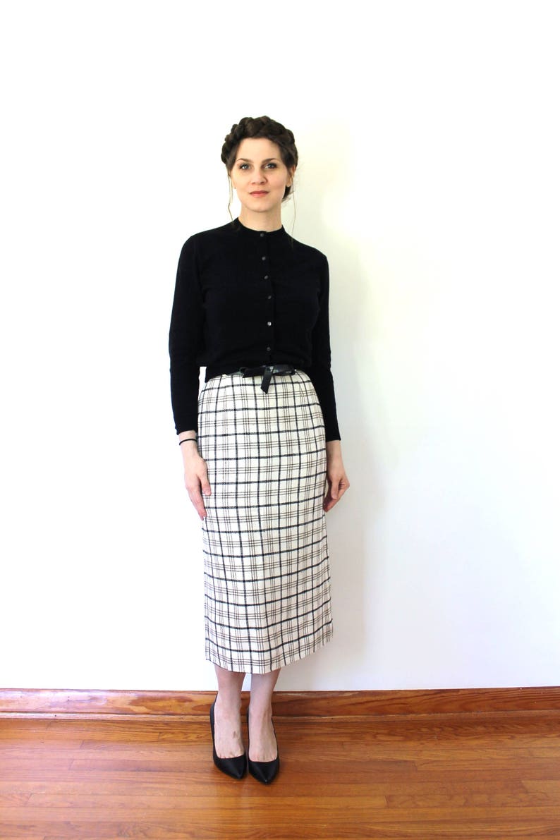 May include: A woman wearing a black cardigan and a white and black plaid skirt. She is wearing black heels.