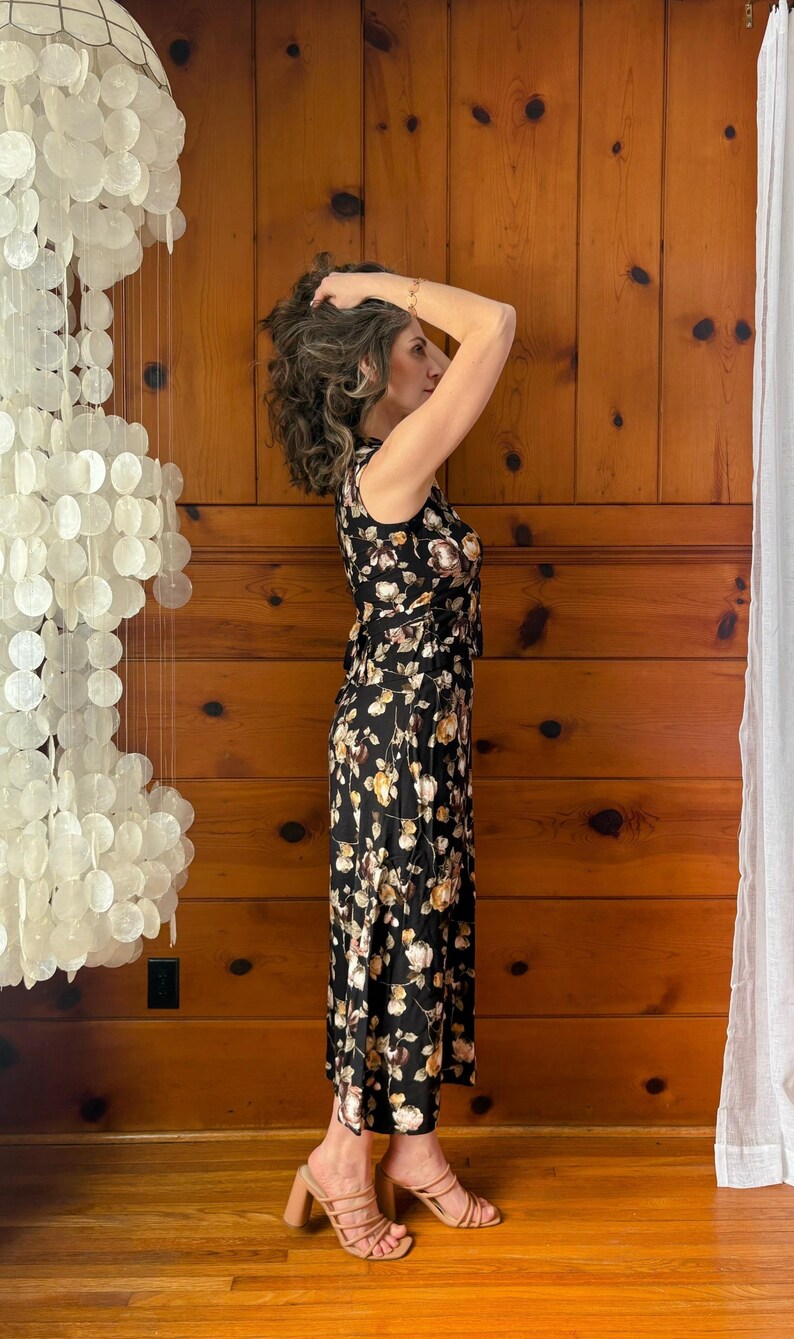 K&ouml;nnte beinhalten: Eine Frau tr&auml;gt ein schwarzes Maxikleid mit Blumenmuster und braune Sandalen mit Absatz. Das Kleid hat ein figurbetontes Oberteil und einen flie&szlig;enden Rock. Die Frau steht vor einer Holzvert&auml;felung.