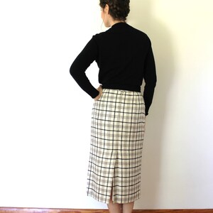 May include: A woman wearing a black mock neck sweater and a white and black plaid skirt. She is standing with her back to the camera and her hands on her hips.