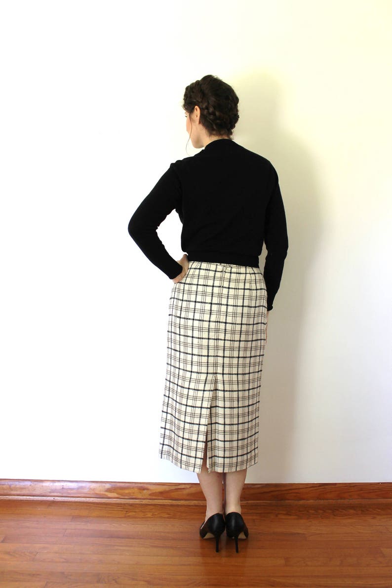 May include: A woman wearing a black mock neck sweater and a white and black plaid skirt. She is standing with her back to the camera and her hands on her hips.