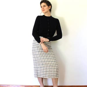 May include: A woman wearing a black cardigan and a white and black plaid skirt. She is standing with her arms crossed and looking to the side. She is wearing black heels.