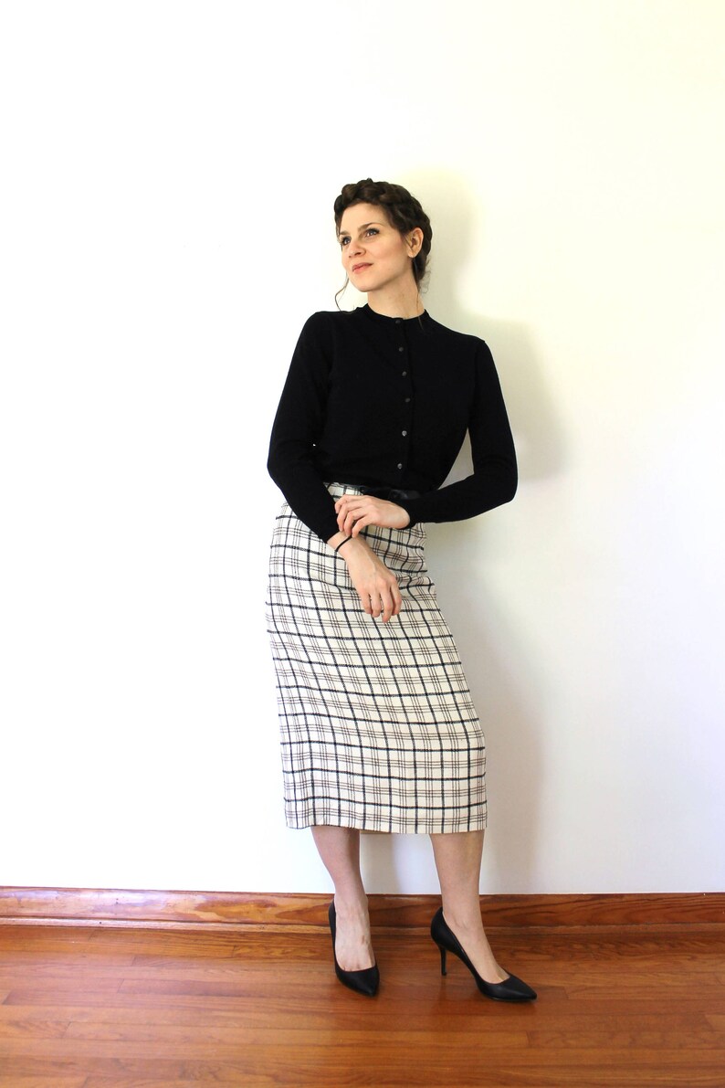 May include: A woman wearing a black cardigan and a white and black plaid skirt. She is standing with her arms crossed and looking to the side. She is wearing black heels.