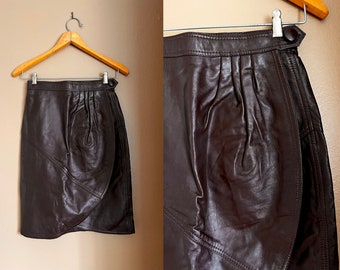 May include: A brown leather skirt with a wrap-around design and a pleated front. The skirt has a zipper closure and a hook and eye closure.