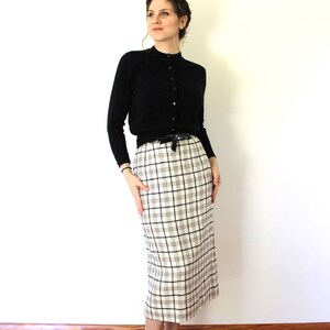 May include: A woman wearing a black cardigan, a white and black plaid skirt, and black heels. The skirt has a black belt with a bow.