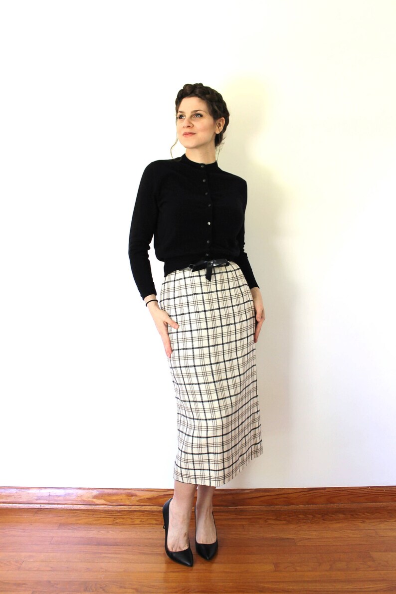 May include: A woman wearing a black cardigan, a white and black plaid skirt, and black heels. The skirt has a black belt with a bow.