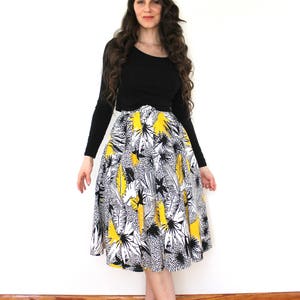 May include: A woman wearing a black long-sleeved shirt and a yellow, black, and white floral print skirt. She is standing on a wooden floor with a white wall in the background.