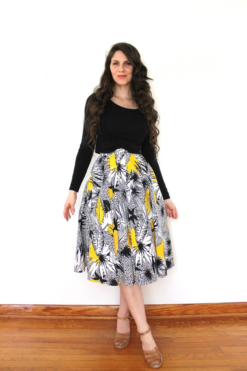 May include: A woman wearing a black long-sleeved shirt and a yellow, black, and white floral print skirt. She is standing on a wooden floor with a white wall in the background.