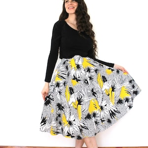 May include: A woman wearing a black long-sleeved shirt and a black and white skirt with a yellow floral print. The skirt is a full skirt and the woman is standing on a wooden floor.