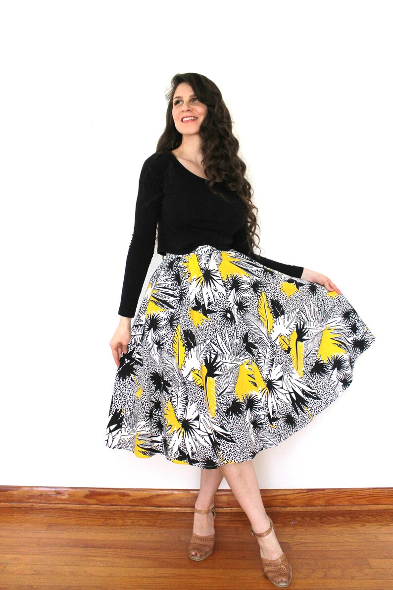 May include: A woman wearing a black long-sleeved shirt and a black and white skirt with a yellow floral print. The skirt is a full skirt and the woman is standing on a wooden floor.