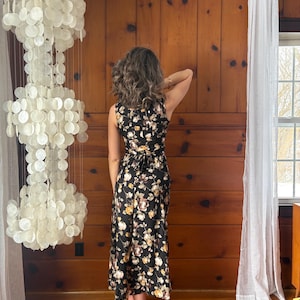 K&ouml;nnte beinhalten: Ein schwarzes Maxikleid mit Blumenmuster. Das Kleid ist &auml;rmellos und hat einen Wickelrock. Das Model tr&auml;gt das Kleid in einem Raum mit Holzvert&auml;felung und einem wei&szlig;en Vorhang.