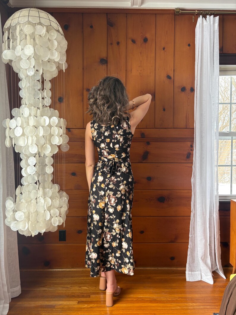 K&ouml;nnte beinhalten: Ein schwarzes Maxikleid mit Blumenmuster. Das Kleid ist &auml;rmellos und hat einen Wickelrock. Das Model tr&auml;gt das Kleid in einem Raum mit Holzvert&auml;felung und einem wei&szlig;en Vorhang.