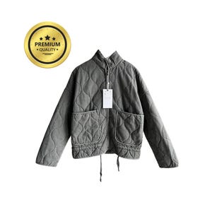 Könnte beinhalten: Gesteppte olivgrüne Jacke mit hohem Kragen, zwei Vordertaschen und einem Kordelzug am Saum. Die Jacke hat einen Reißverschluss und ein Etikett. Ein goldenes Siegel mit "PREMIUM QUALITY" befindet sich in der oberen linken Ecke.