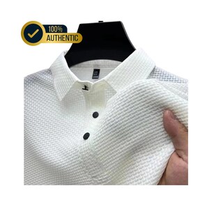 May include: White textured polo shirt with black buttons and a collar. The shirt is displayed on a black hanger. A gold and black badge in the upper left corner reads "100% AUTHENTIC".
