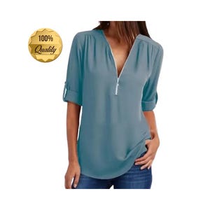 May include: A teal-colored blouse with a V-neck and a silver zipper. The sleeves are rolled up, and the blouse has a loose fit. A gold badge with the text "100% Quality" is in the upper left corner.