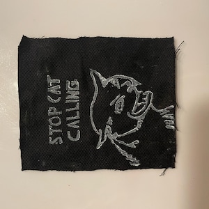 May include: Black fabric patch with white embroidered text that reads "STOP CAT CALLING" and a white embroidered cartoon cat with a surprised expression.