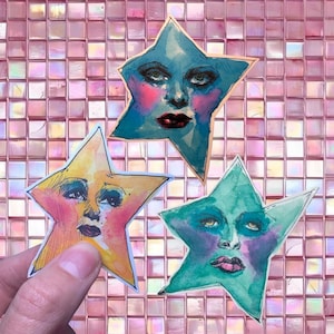 Puede incluir: Tres obras de arte en acuarela con forma de estrella y rostros humanos. Las estrellas están pintadas en tonos de azul, amarillo y verde, con rubor rosa y labios rojo oscuro. Las obras de arte se sostienen sobre un fondo rosa con baldosas cuadradas.