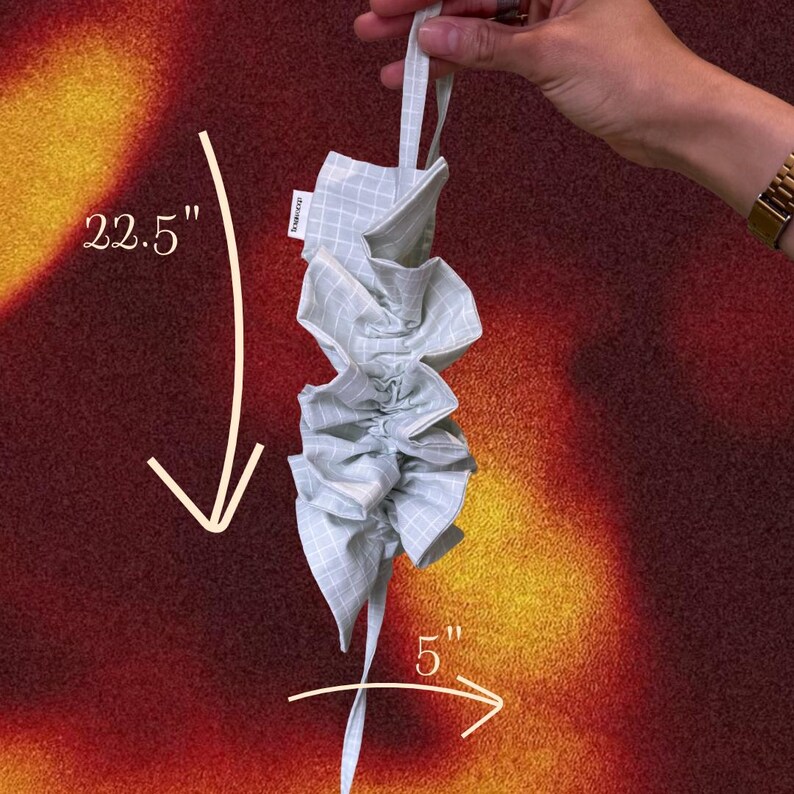 May include: A light green and white checkered fabric item, approximately 22.5 inches long and 5 inches wide, is held up by a loop. The fabric is gathered and ruffled, with a tag that reads "KOKOSON".