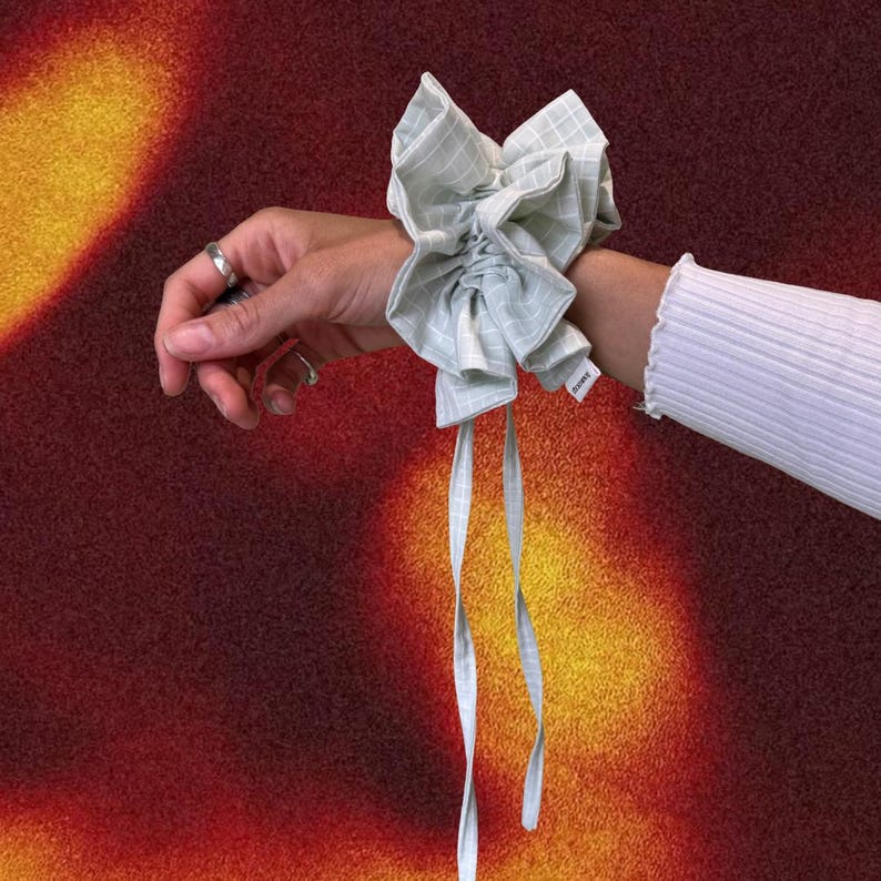 May include: A light green and white checkered scrunchie with long, matching ribbons tied around a wrist. The scrunchie is made of a soft fabric and is gathered to create a voluminous effect. The background is a gradient of red, orange, and yellow.