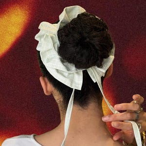 May include: A light green and white checkered fabric head covering tied in a bow. The head covering is wrapped around a bun of dark hair. The background is a gradient of red and yellow.