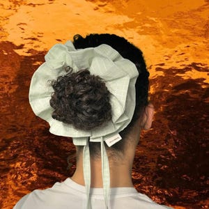 May include: A light green and white checkered bonnet with a dark brown bun. The bonnet has long ties and a small tag. The background is a textured orange and gold.