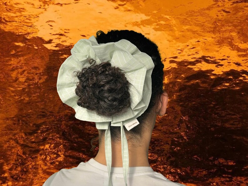 May include: A light green and white checkered bonnet with a dark brown bun. The bonnet has long ties and a small tag. The background is a textured orange and gold.