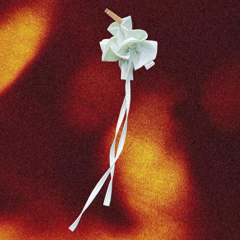 May include: A light green fabric item, possibly a sachet or decorative element, is held by a wooden clothespin. Long, thin white ribbons extend from the fabric. The background is a textured blend of red, orange, and yellow.
