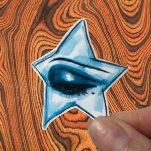 Può includere: Un adesivo blu a forma di stella con un'illustrazione dettagliata di un occhio chiuso. L'occhio è ombreggiato in blu scuro, con un contorno blu chiaro. L'adesivo è tenuto su uno sfondo di venature del legno.