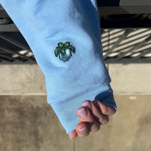 Puede incluir: Manga de sudadera azul claro con un diseño pixelado verde y azul cerca del puño. El puño y el diseño son de un tono azul más oscuro. La manga está hecha de un material suave.