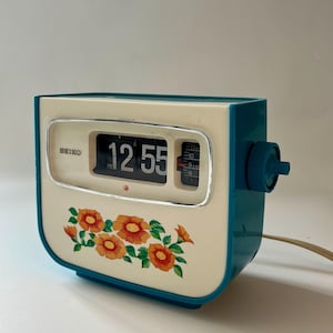 May include: A vintage Seiko flip clock with a teal blue outer casing and a cream-coloured front panel. The face features a floral pattern of orange flowers with green leaves, a digital flip display showing 12:55, and a side adjustment knob.
