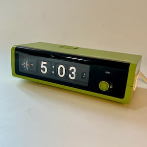 Peut inclure: Une horloge à bascule numérique de style rétro, de couleur verte vive. L'horloge affiche l'heure 5:03 en chiffres blancs sur fond noir. Elle a un design rectangulaire épuré avec un petit bouton et un cordon d'alimentation.