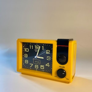 May include: A vintage yellow Citizen quartz alarm clock with a black face. The clock displays the time as 10:35. It has a speaker and a volume control knob. The clock is rectangular with rounded corners.