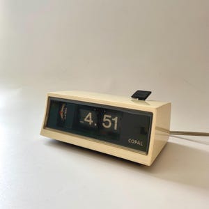 May include: A vintage cream-colored Copal flip clock displaying 4:51 PM. The clock has a black top button and a green and black display. The word "Copal" is printed on the front. A power cord extends from the side.
