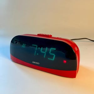May include: A red and black Seiko digital alarm clock showing 7:45 AM in green numerals. The clock has a curved design and a small red button on top. The Seiko logo is visible on the front.