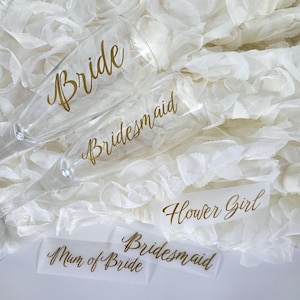 May include: Clear champagne flutes with gold lettering that reads "Bride" and "Bridesmaid." Also, there are labels that say "Flower Girl," "Bridesmaid," and "Mum of Bride." The items are on a white, textured surface.