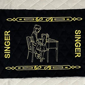 May include: Black fabric with gold embroidery of a woman sitting at a sewing machine. The fabric has the word "SINGER" embroidered on each side.