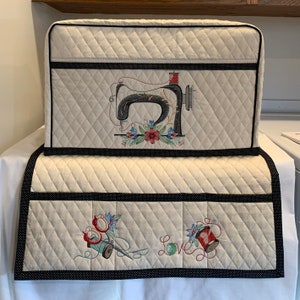 May include: A cream-colored quilted sewing machine cover and matching mat featuring black polka-dot trim. The cover displays an embroidered vintage sewing machine with floral accents, while the mat features embroidered scissors, thread, and the word Love.