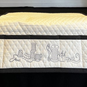 May include: White quilted fabric storage organizer with black polka dot trim. The organizer has a pocket with an embroidered design of sewing supplies, including spools of thread, needles, and a thimble.