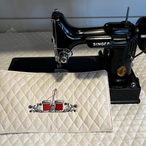 May include: A black Singer sewing machine with a white quilted sewing mat with an embroidered design of two spools of thread and a needle. The mat has a diamond pattern.