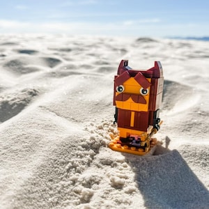 Op de afbeelding: Een Lego-figuur van een personage met een bruine snor, rode oren en een gele gezicht en torso. De figuur staat op een zanderige ondergrond onder een blauwe lucht.