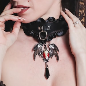 May include: A black choker necklace with a silver bat pendant featuring a red gemstone. The pendant has a skull design and a black crystal hanging from the bottom.