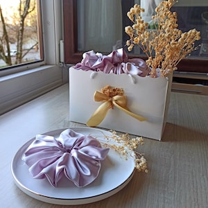 Peut inclure: Un chouchou en satin lilas sur une assiette blanche à bord doré. Un sac cadeau blanc avec un nœud doré et des fleurs séchées contient d'autres chouchous. Des fleurs séchées sont également à côté de l'assiette. Les articles sont sur une surface en bois.
